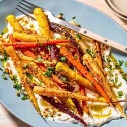 Roasted rainbow carrots with with dukkah, mint and a garlicky yogurt sauce on a serving platter with a fork. Next to this is a small bowl with chopped mint and a stack of 2 plates.