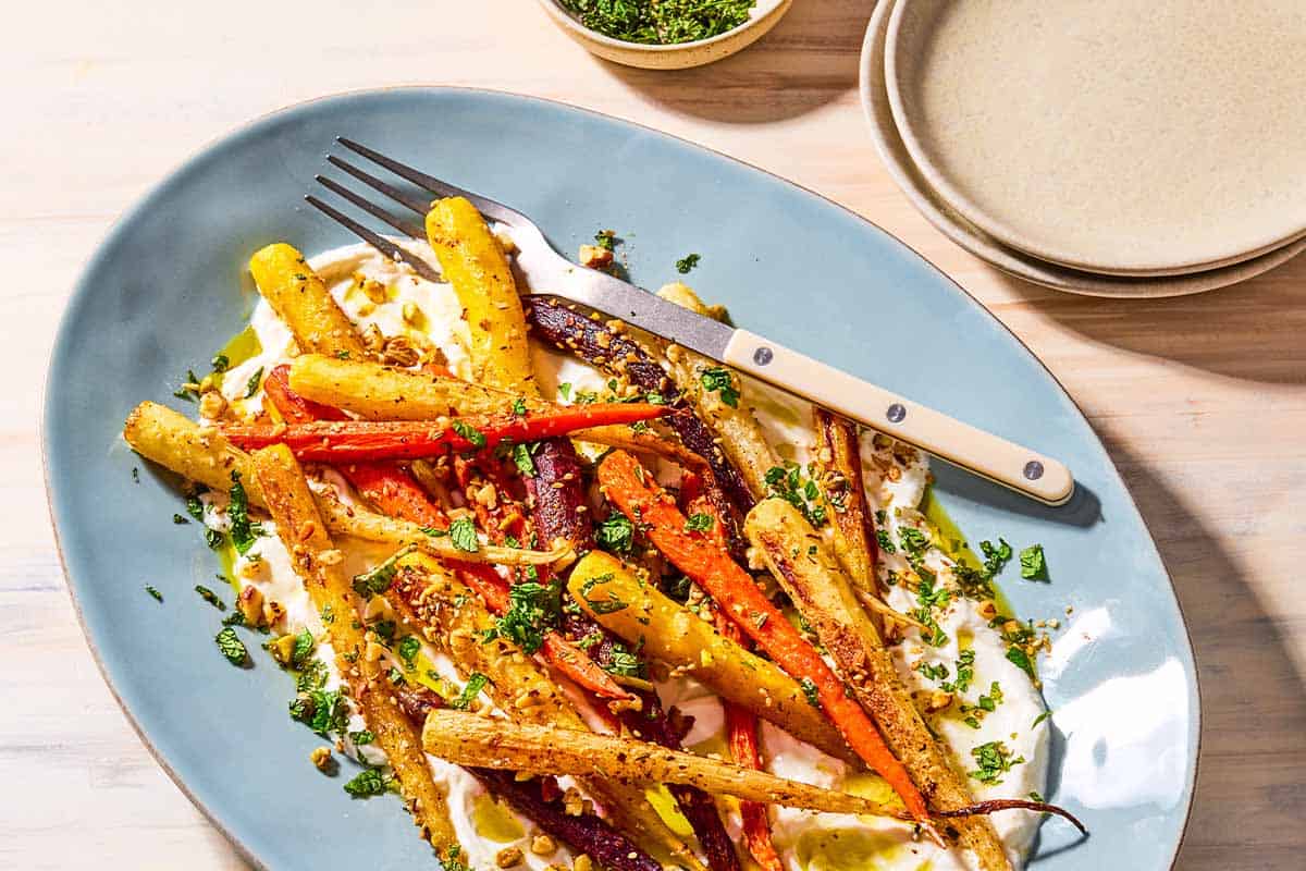 An overhead close up photo of roasted rainbow carrots with with dukkah, mint and a garlicky yogurt sauce on a serving platter with a fork. Next to this is a small bowl with chopped mint and a stack of 2 plates.