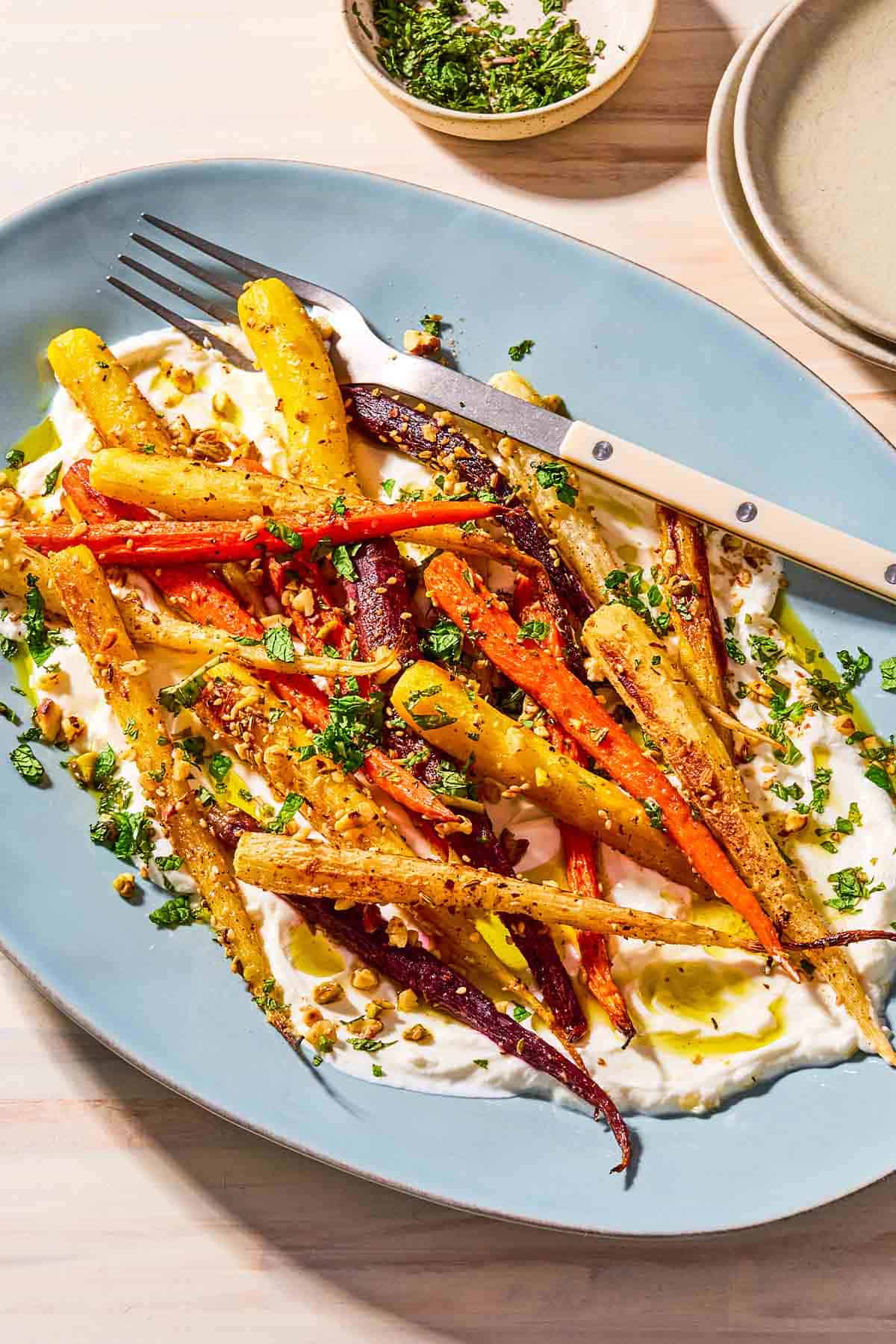 Roasted rainbow carrots with with dukkah, mint and a garlicky yogurt sauce on a serving platter with a fork. Next to this is a small bowl with chopped mint and a stack of 2 plates.
