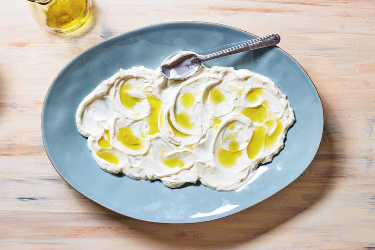 The garlicky yogurt topped with a drizzle of olive oil on a serving platter with a spoon. Next to this is a cup of olive oil.