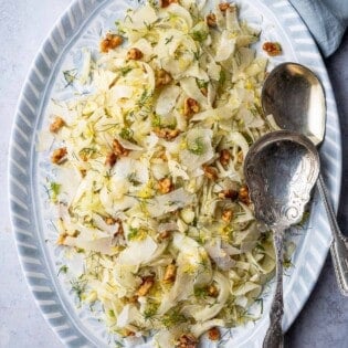 Overhead shot of fennel salad on a blue and white serving platter with two silver serving spoons.