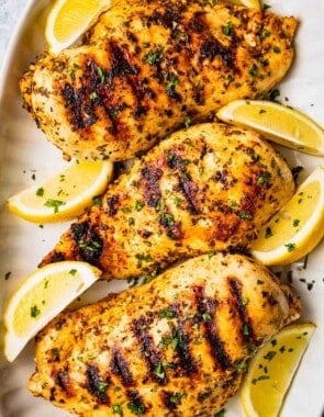 Three grilled chicken breasts that have been marinated in Greek chicken marinade and are on a platter with lemon wedges and a sprinkle of parsley.