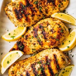 Three grilled chicken breasts that have been marinated in Greek chicken marinade and are on a platter with lemon wedges and a sprinkle of parsley.