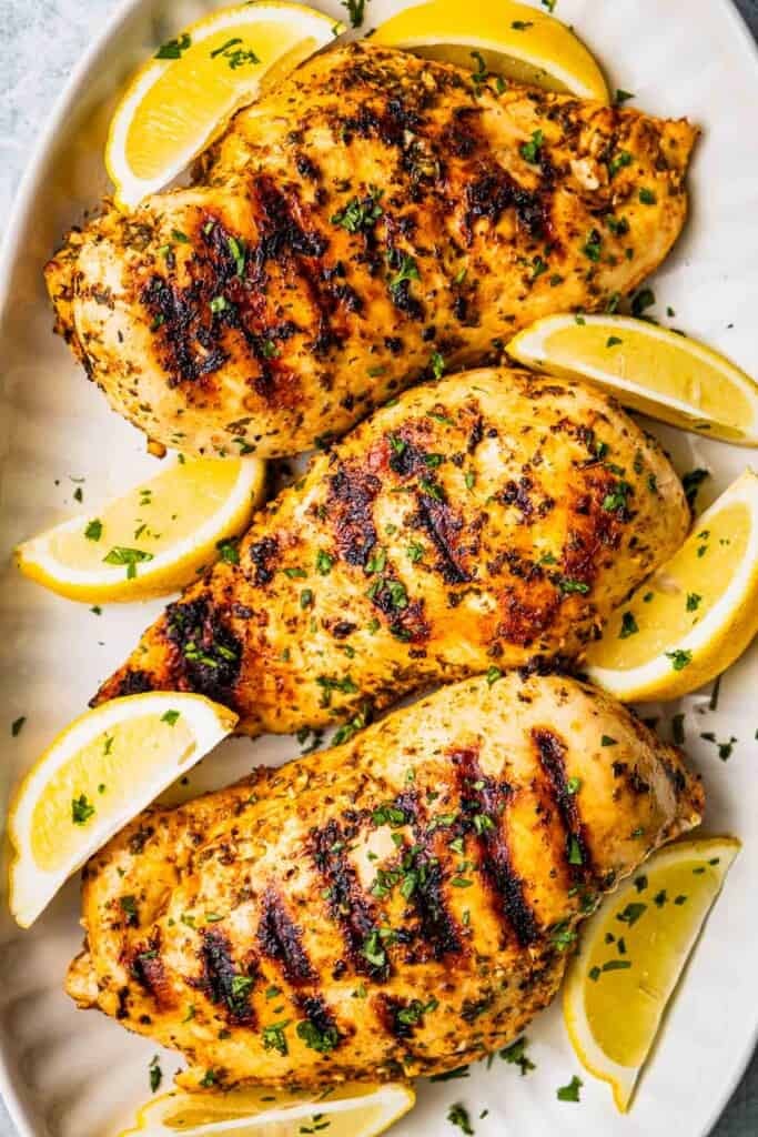 Three grilled chicken breasts that have been marinated in Greek chicken marinade and are on a platter with lemon wedges and a sprinkle of parsley.