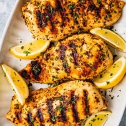 Overhead shot of three grilled chicken breasts that have been marinated in Greek chicken marinade and are on a platter with lemon wedges and a sprinkle of parsley.