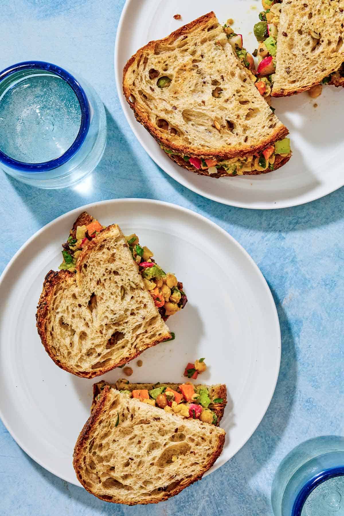 An overhead photo of two plates, each containing 2 vegan chickpea salad sandwich halves, next to two glasses of water.