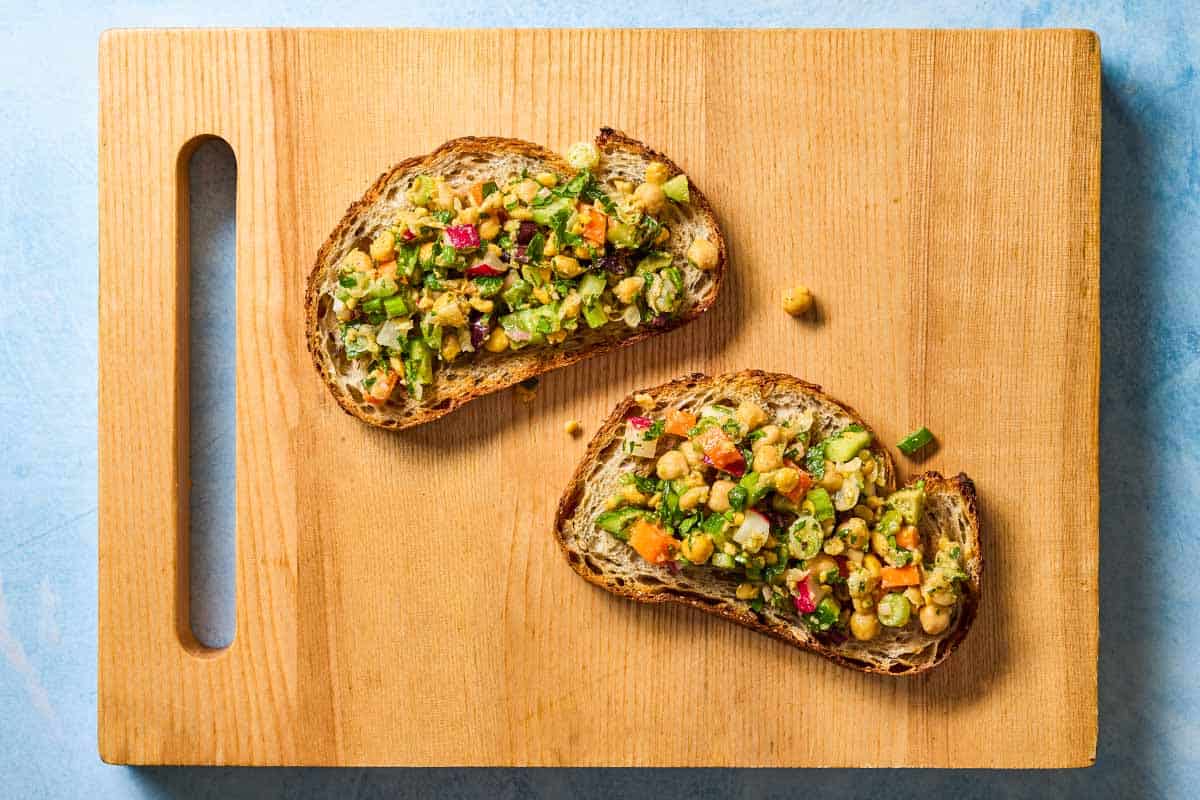 two slices of toast on a wooden cutting board, each topped with vegan chickpea salad.