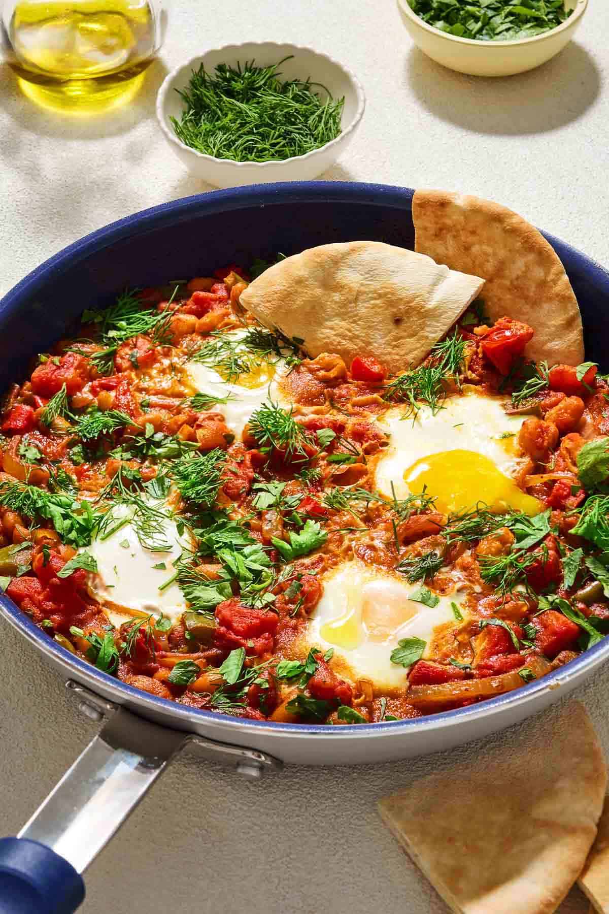 White bean shakshuka garnished with dill and parsley in a skillet with 2 pieces of pita bread. Next to this are small bowls of dill and parsley, a cup of olive oil, and more pieces of pita bread.