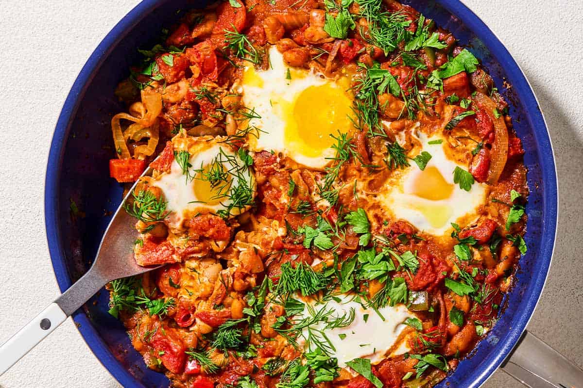 An overhead photo of white bean shakshuka garnished with dill and parsley in a skillet with a serving spoon.