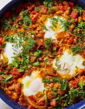 An overhead photo of white bean shakshuka garnished with dill and parsley in a skillet. Next to this are small bowls of dill and parsley.