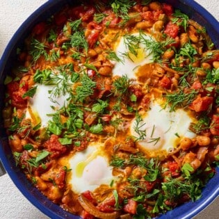 An overhead photo of white bean shakshuka garnished with dill and parsley in a skillet. Next to this are small bowls of dill and parsley.