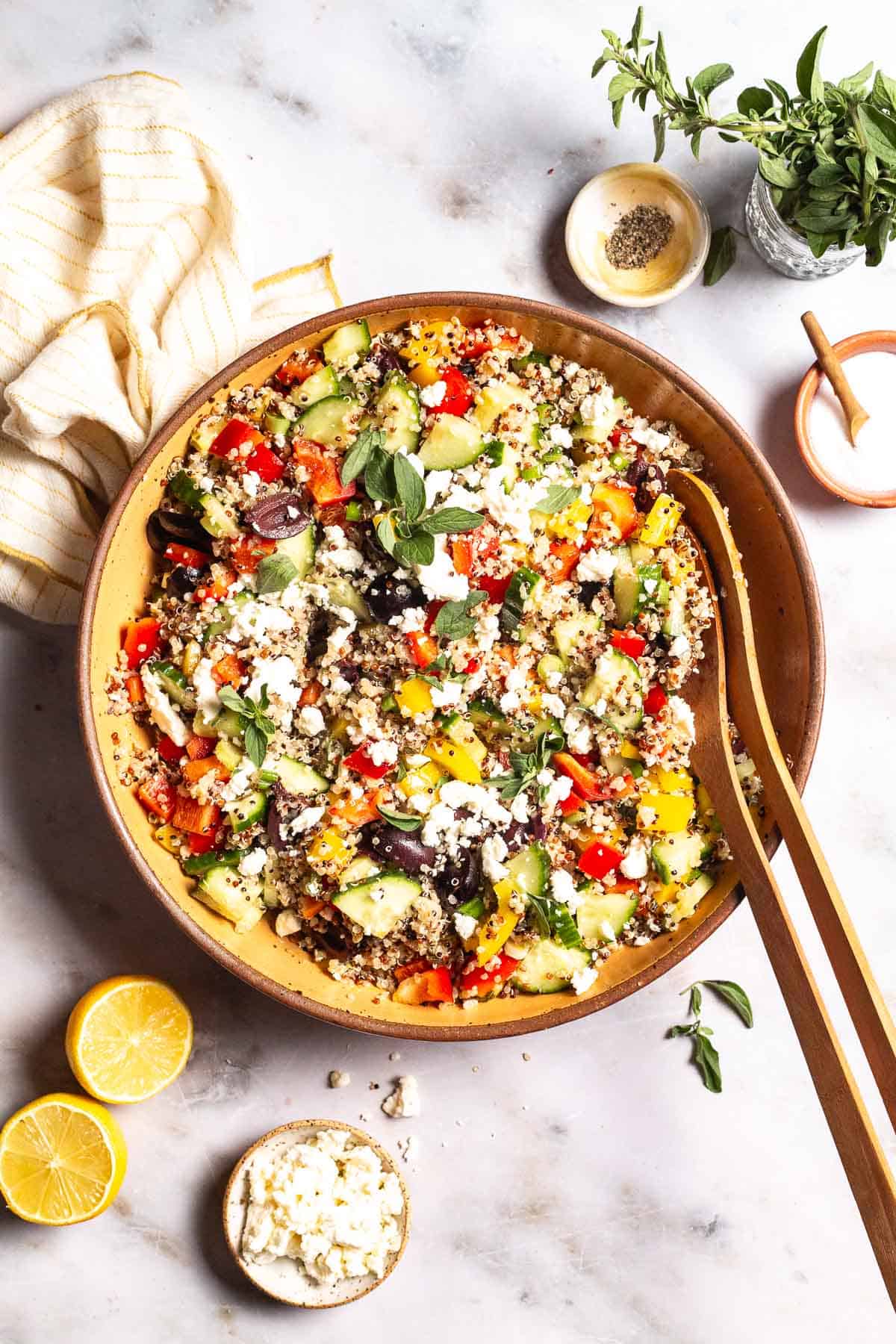 An overhead photo of quinoa salad in a large bowl with wooden serving utensils. This is surrounded by small bowls of feta cheese, salt and pepper as well as 2 lemon halves, a jar of fresh oregano, and a cloth napkin.