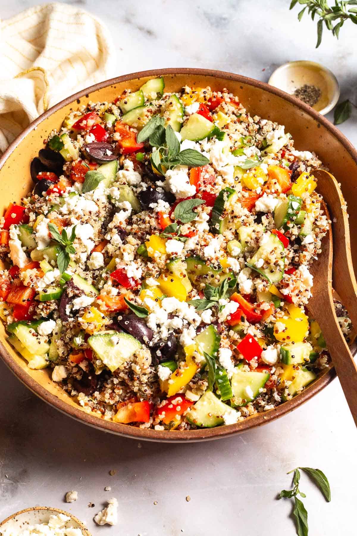 Quinoa salad in a large bowl with wooden serving utensils. This is surrounded by small bowls of feta cheese and pepper and a cloth napkin.