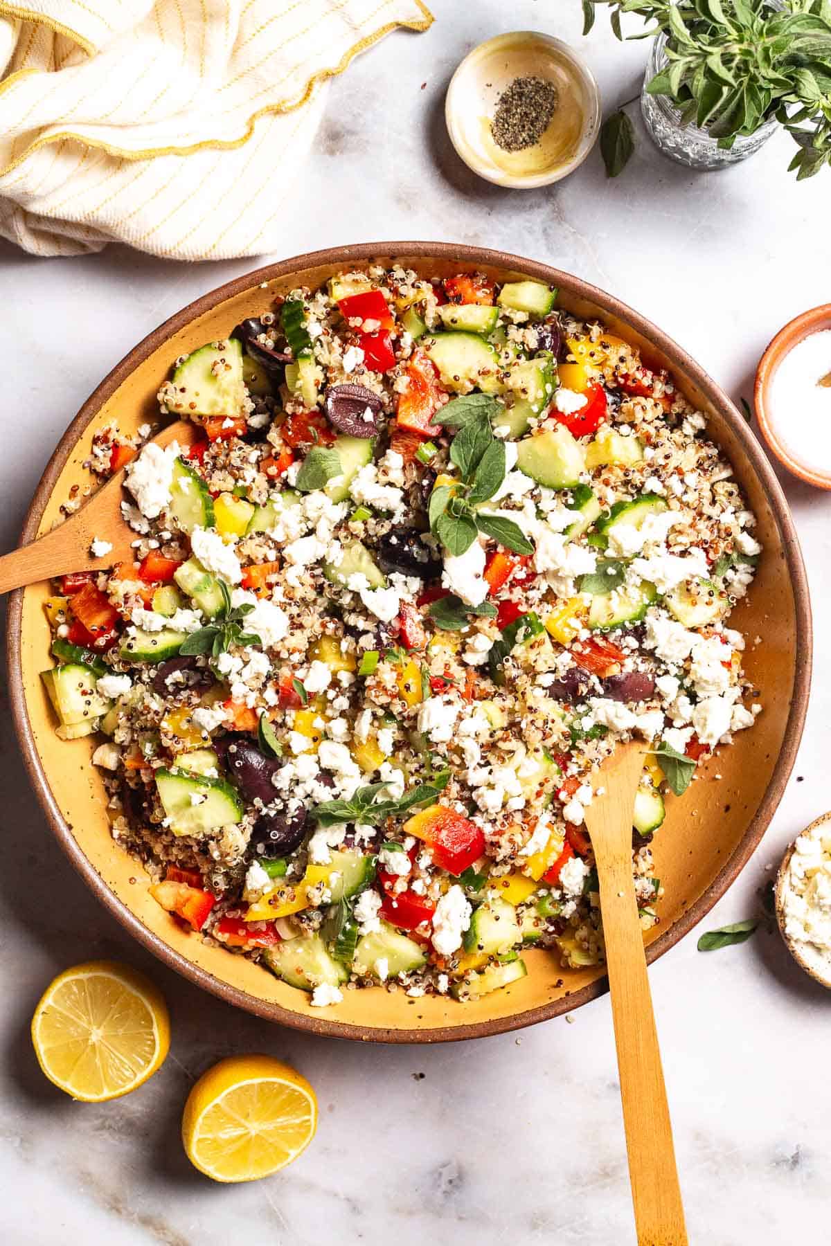 An overhead photo of quinoa salad in a large bowl with wooden serving utensils. This is surrounded by small bowls of feta cheese, salt and pepper as well as 2 lemon halves, a jar of fresh oregano, and a cloth napkin.