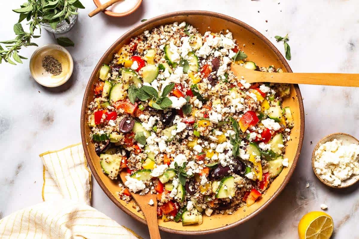 Quinoa salad in a large bowl with wooden serving utensils. This is surrounded by small bowls of feta cheese, salt and pepper as well as a lemon half, a jar of fresh oregano, and a cloth napkin.