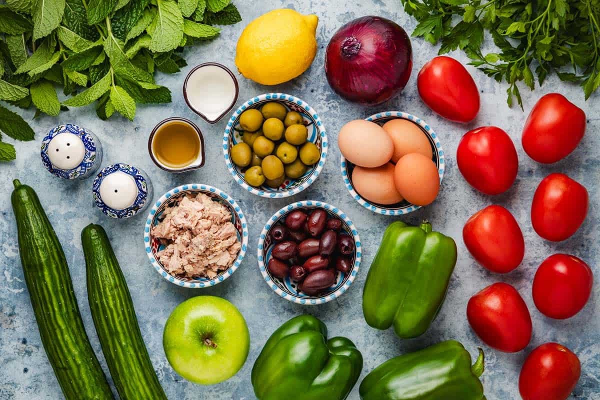 ingredients for tunisian salad including tomatoes, red onion, eggs, green peppers, a green apple, green olives, kalamata olives, lemon, olive oil, white wine vinegar, tuna, salt, pepper, english cucumbers, mint and parsley.