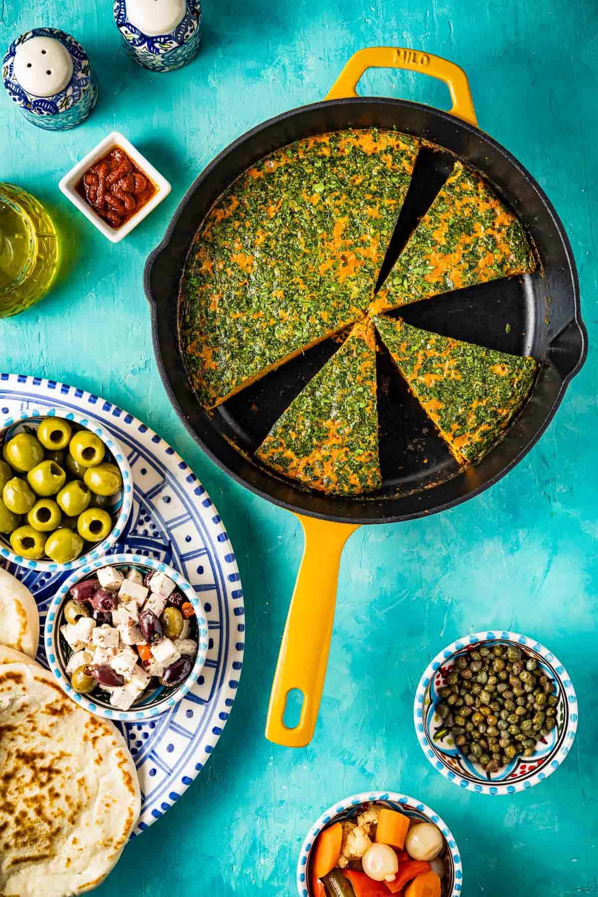 An overhead photo of Tunisian style baked frittata with carrot cut into slices in a skillet. This is surrounded pita bread, salt and pepper shakers, and small bowls of harissa, capers, green olives, marinated feta and olives, and giadiniera.