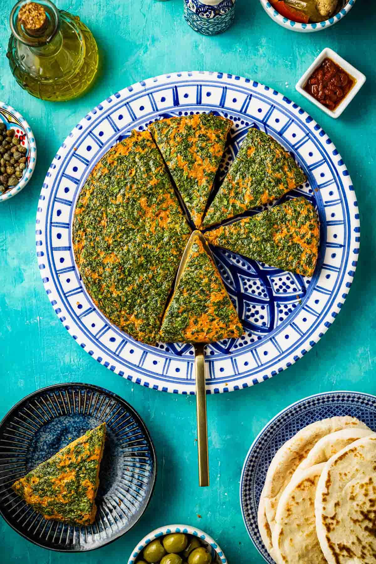 Tunisian style baked frittata with carrot cut into slices on a serving platter with a serving utensil. This is surrounded by a slice of the frittata on a plate, some pita bread, olive oil, and small bowls of harissa, capers and green olives.