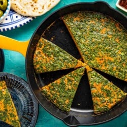 Tunisian style baked frittata with carrot cut into slices in a skillet. This is surrounded by 2 slices of the frittata on two plates, some pita bread, and small bowls of harissa, capers and green olives.