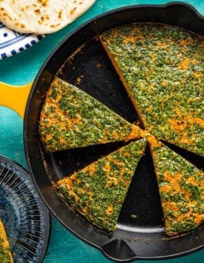 Tunisian style baked frittata with carrot cut into slices in a skillet. This is surrounded by 2 slices of the frittata on two plates, some pita bread, and small bowls of harissa, capers and green olives.