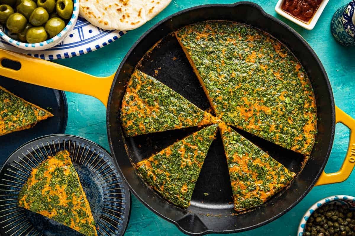 Tunisian style baked frittata with carrot cut into slices in a skillet. This is surrounded by 2 slices of the frittata on two plates, some pita bread, and small bowls of harissa, capers and green olives.