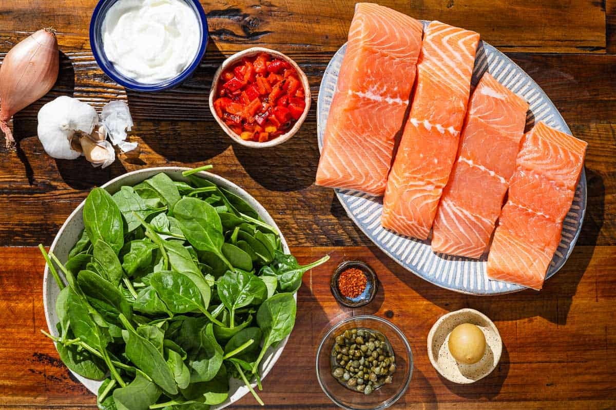 Ingredients for stuffed salmon including salmon fillets, a shallot, garlic, baby spinach, roasted red peppers, capers, greek yogurt, Aleppo pepper, and lemon.