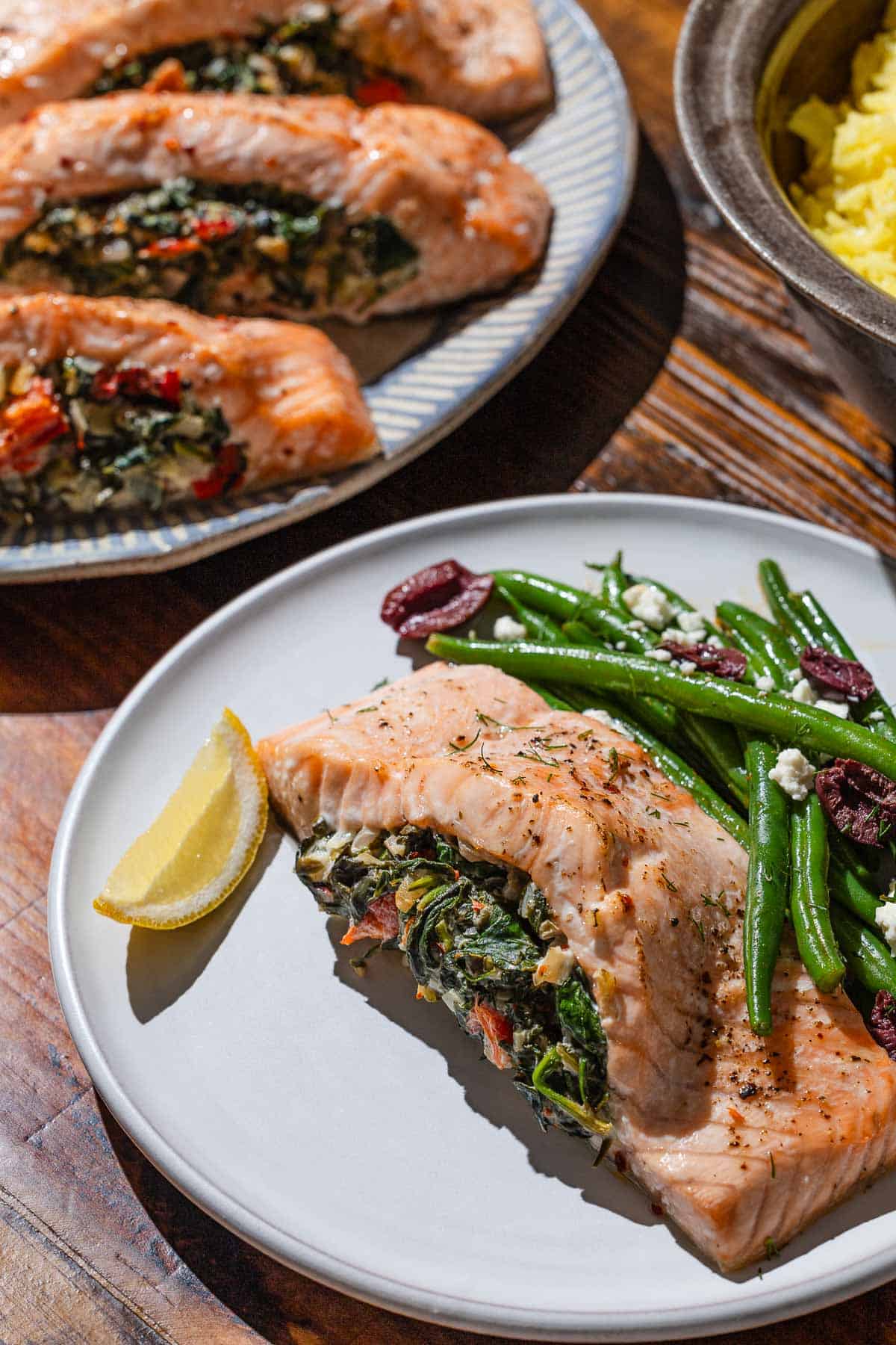 A stuffed salmon fillet on a plate with a serving of green beans, and a lemon wedge. This is next to a serving platter with the rest of the stuffed salmon and a bowl of rice.