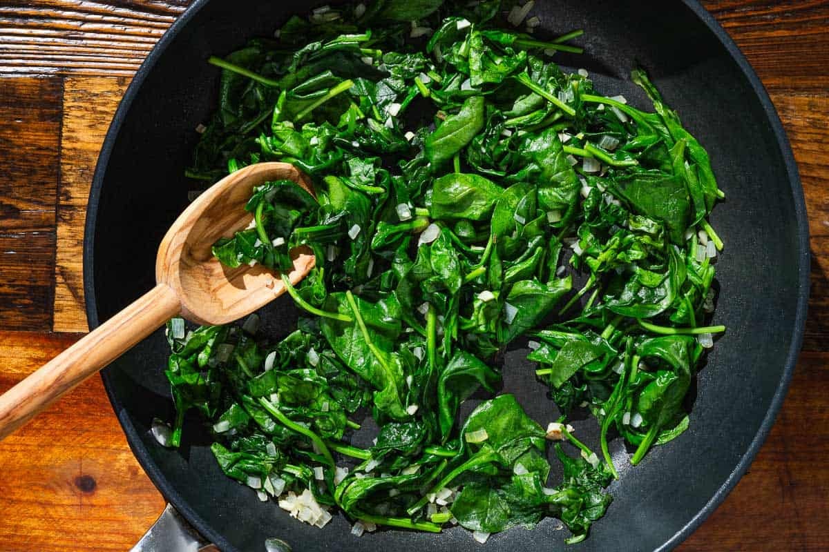 Baby spinach, chopped shallot and garlic sauteeing in a frying pan with a wooden spoon.