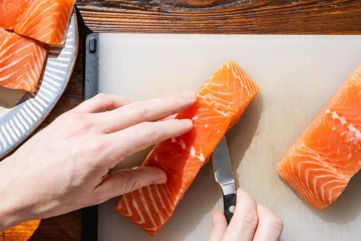 A pocket for the spinach filling being created in a salmon fillet with a knife, next to another fillet on the cutting board, and a platter containing two other salmon fillets.