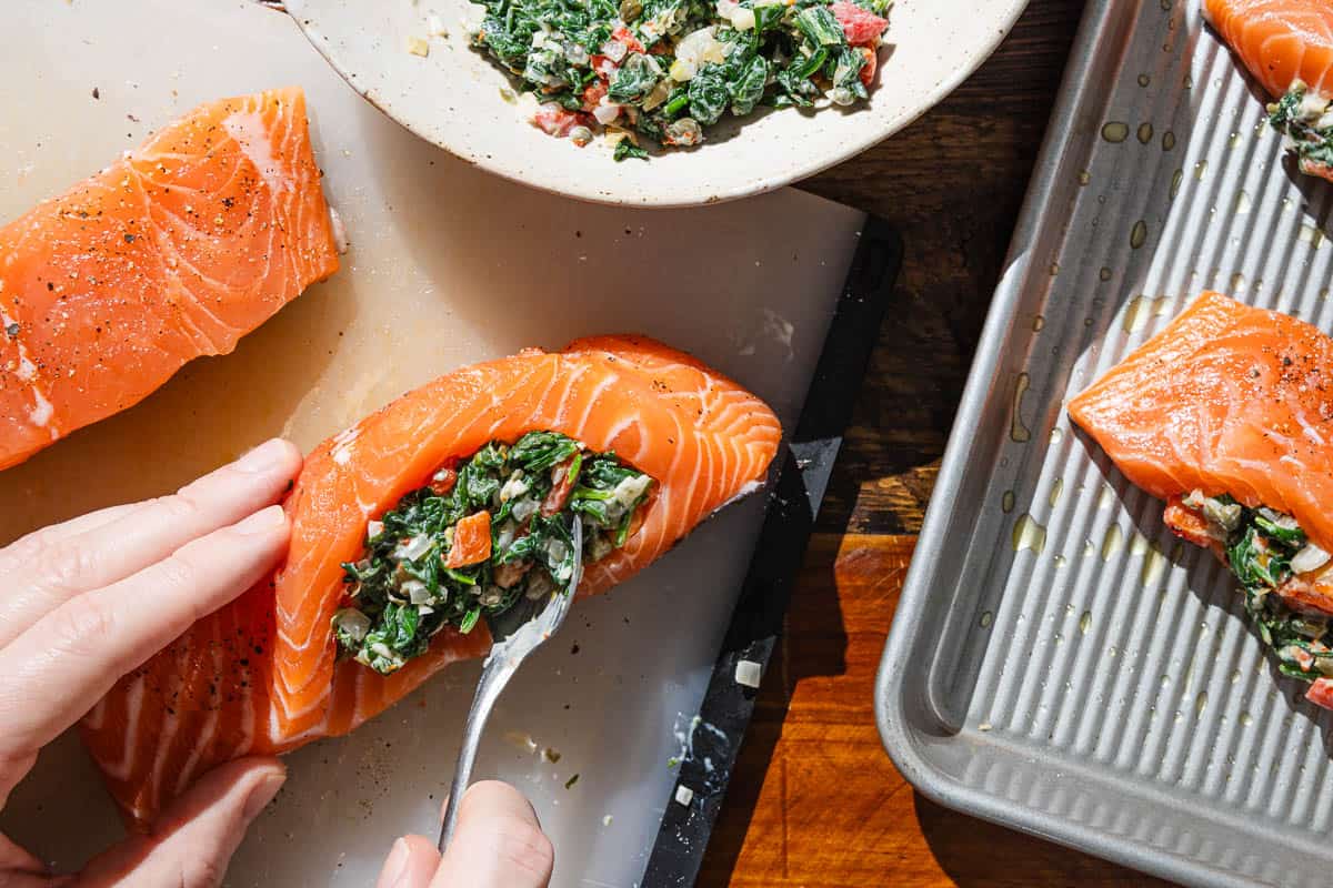 An uncooked salmon fillet being stuffed with a spinach filling, next to a bowl of the filling and a baking sheet with the rest of the stuffed salmon.