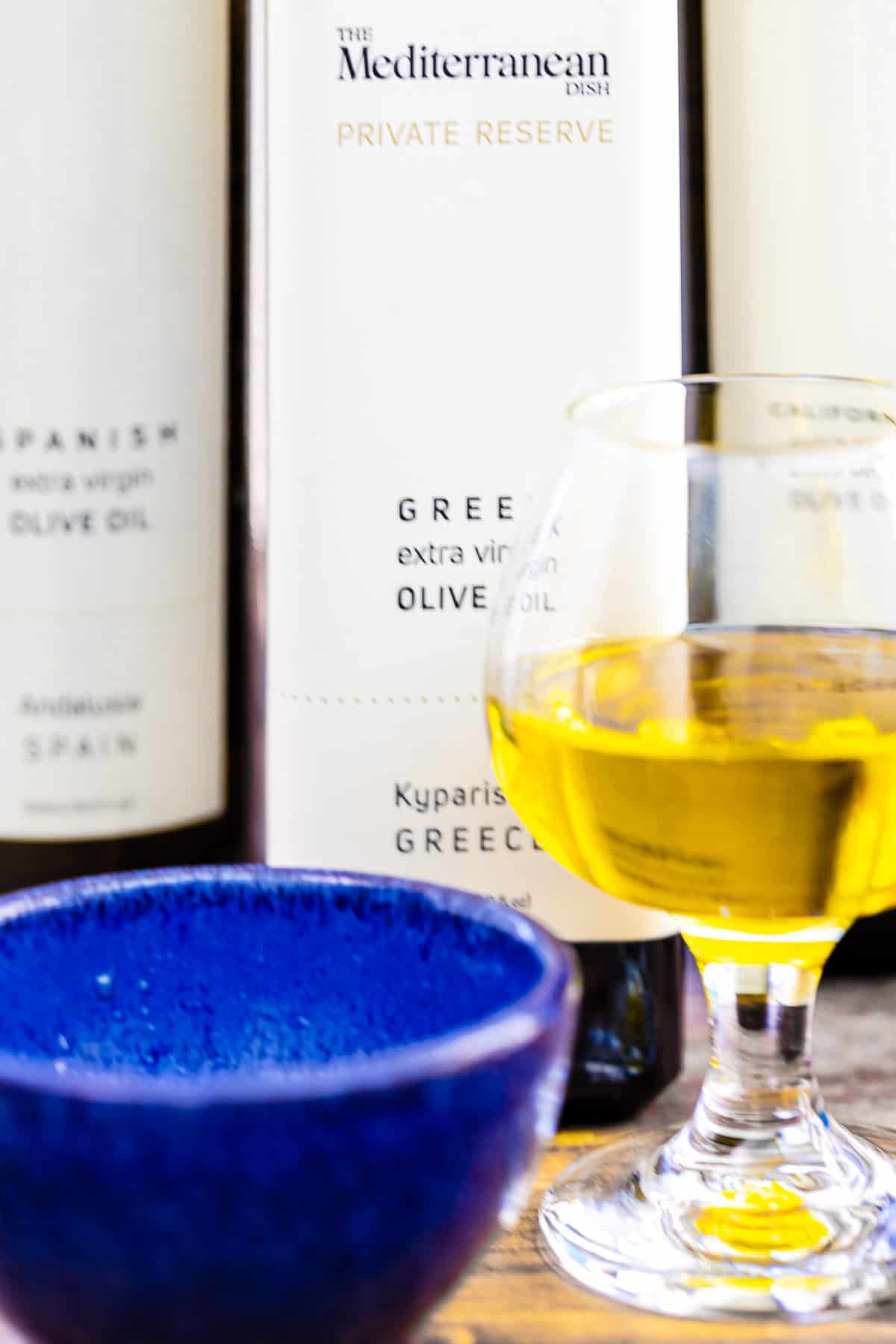 a close up of a small glass of olive oil and a small blue bowl in front of 3 bottles of olive oil.