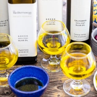 5 different types of olive oils in their bottles sitting behind a half of an apple, a plate of green olives, a small bowl of kalamata olives, and three small glasses and a small blue bowl containing olive oil.