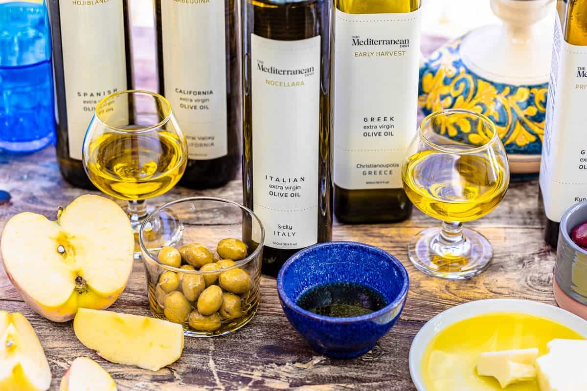 5 different types of olive oils in their bottles sitting behind a half of an apple, apple wedges, a bowl of green olives, a block of cheese in olive oil, and two small glasses and a small blue bowl containing olive oil.
