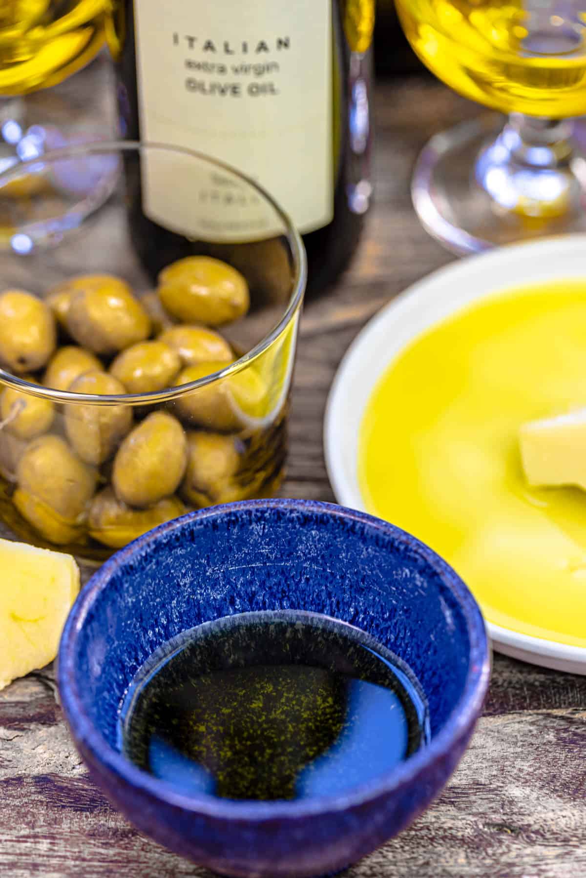 a close up of a small bowl of olive oil next to a bowl of green olives, a plate with olive oil, two small glasses containing olive oil and a bottle of olive oil.