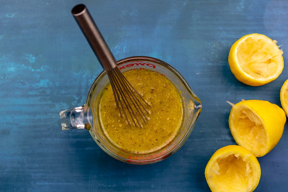 an overhead photo of ladolemono salad dressing in a measuring cup with a whisk next to 3 juiced lemon halves.