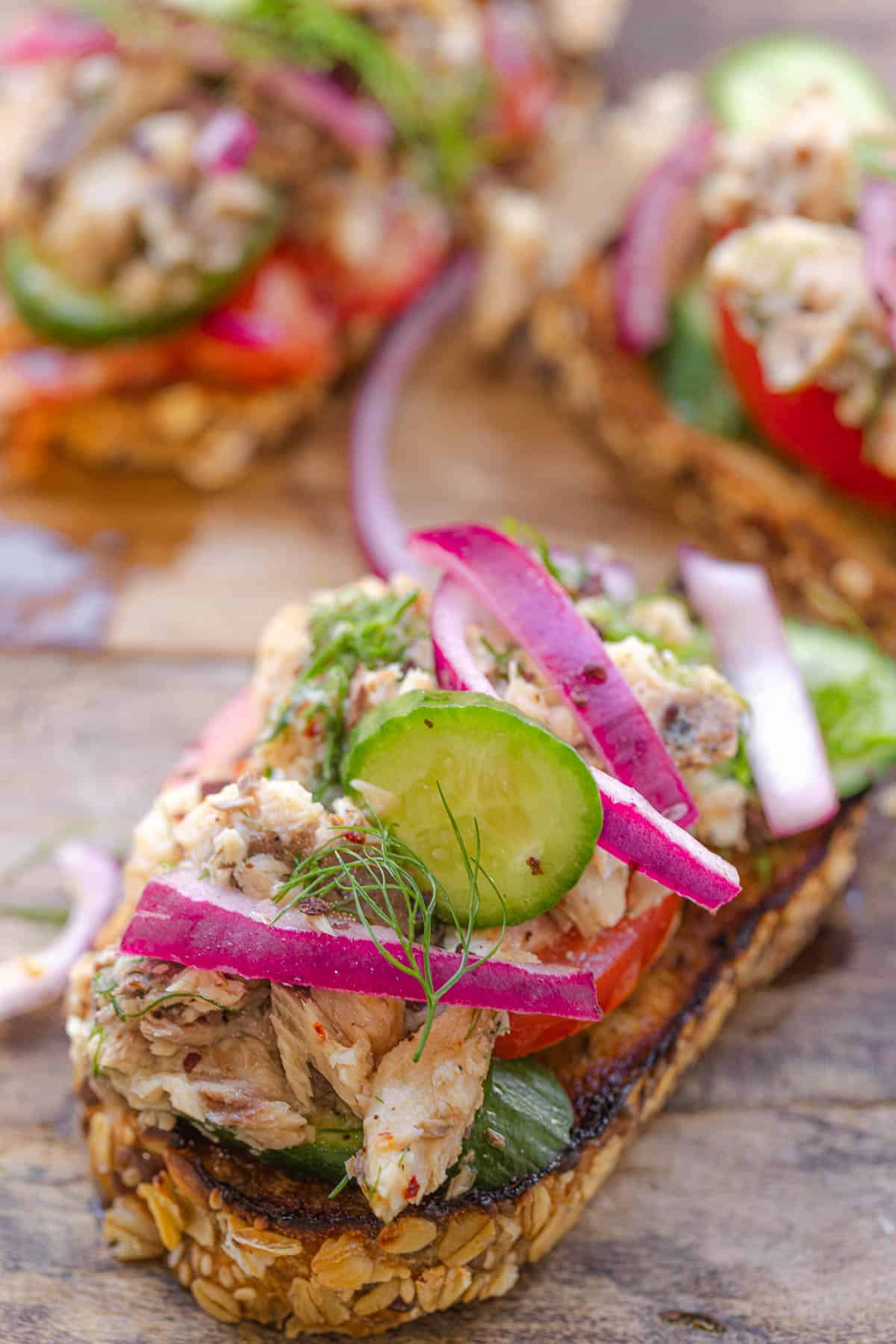 a close up of a sardine toast topped with cucumbers and pickled red onions, in front of two other sardine toasts.