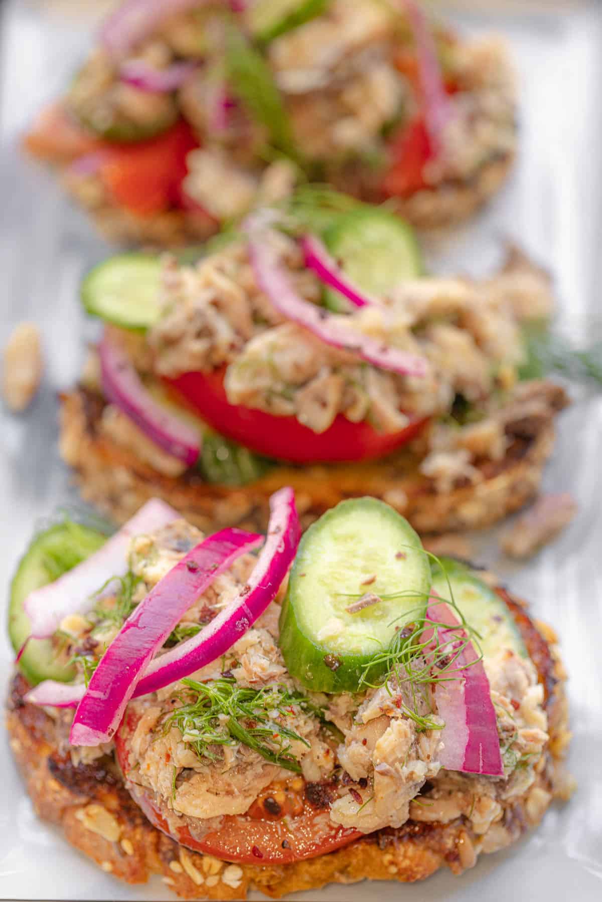 a close up of a sardine toast topped with cucumbers and pickled red onions, in front of two other sardine toasts.