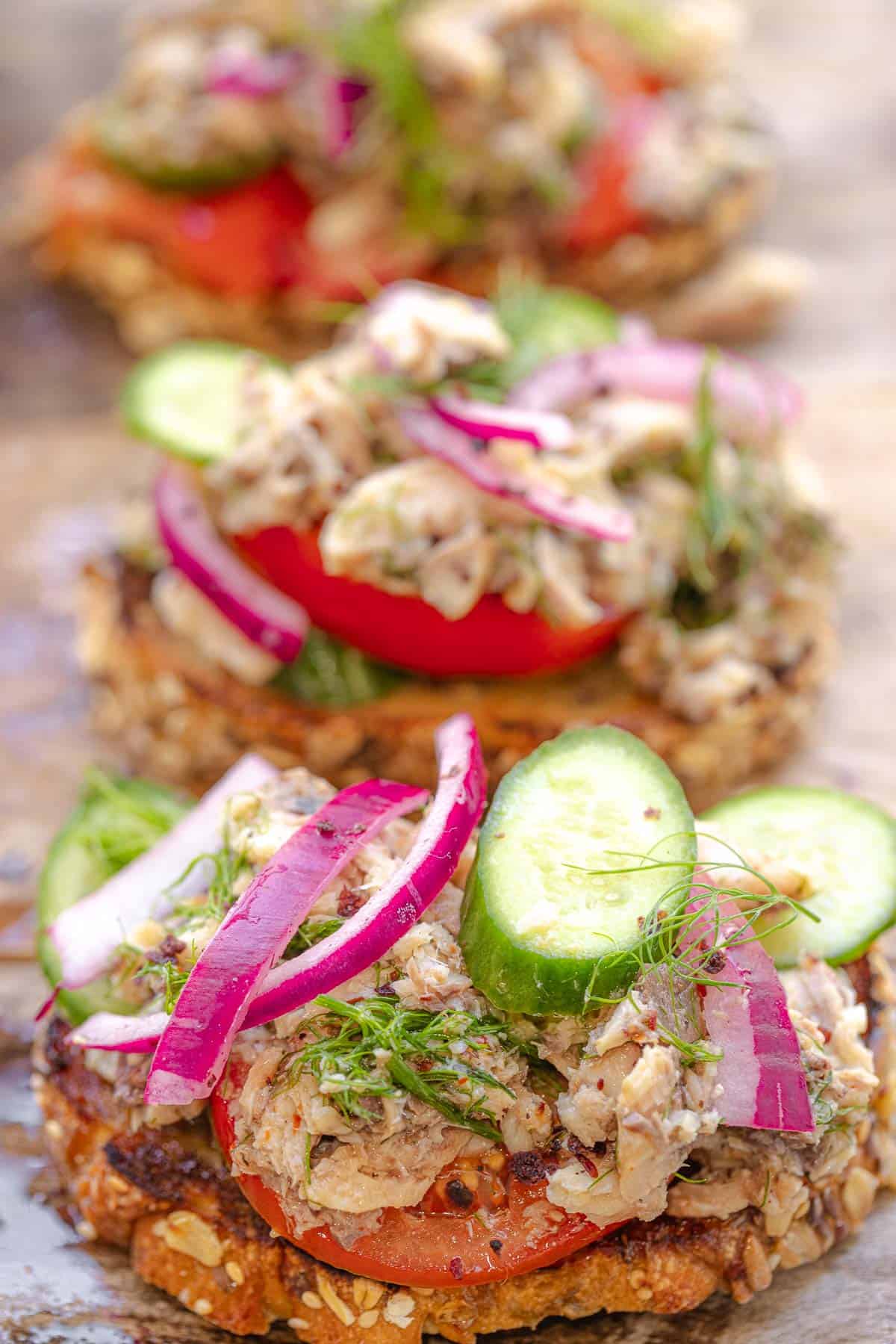 a close up of a sardine toast topped with cucumbers and pickled red onions, in front of two other sardine toasts.