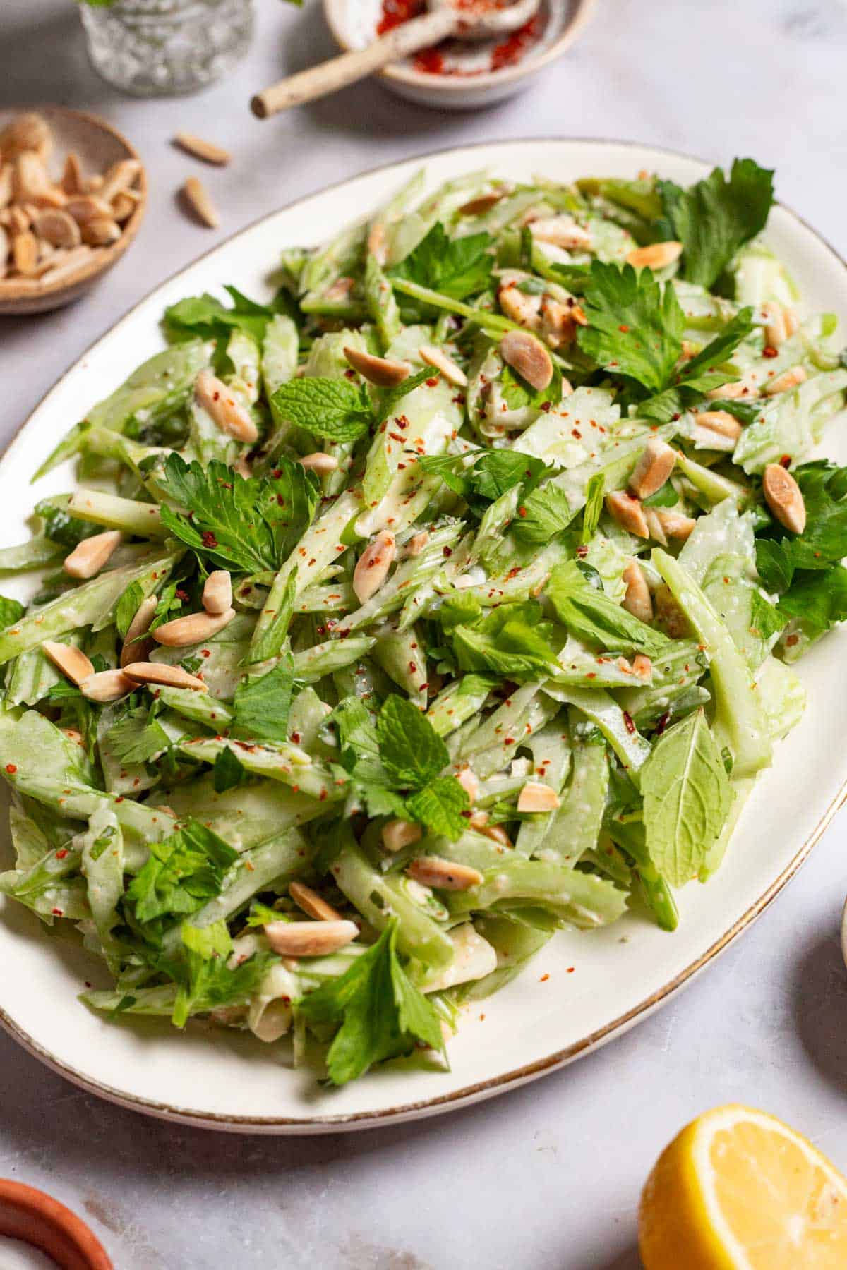 A close up of celery salad on a serving platter. Next to this are bowls of slivered almonds, aleppo pepper and a lemon half.