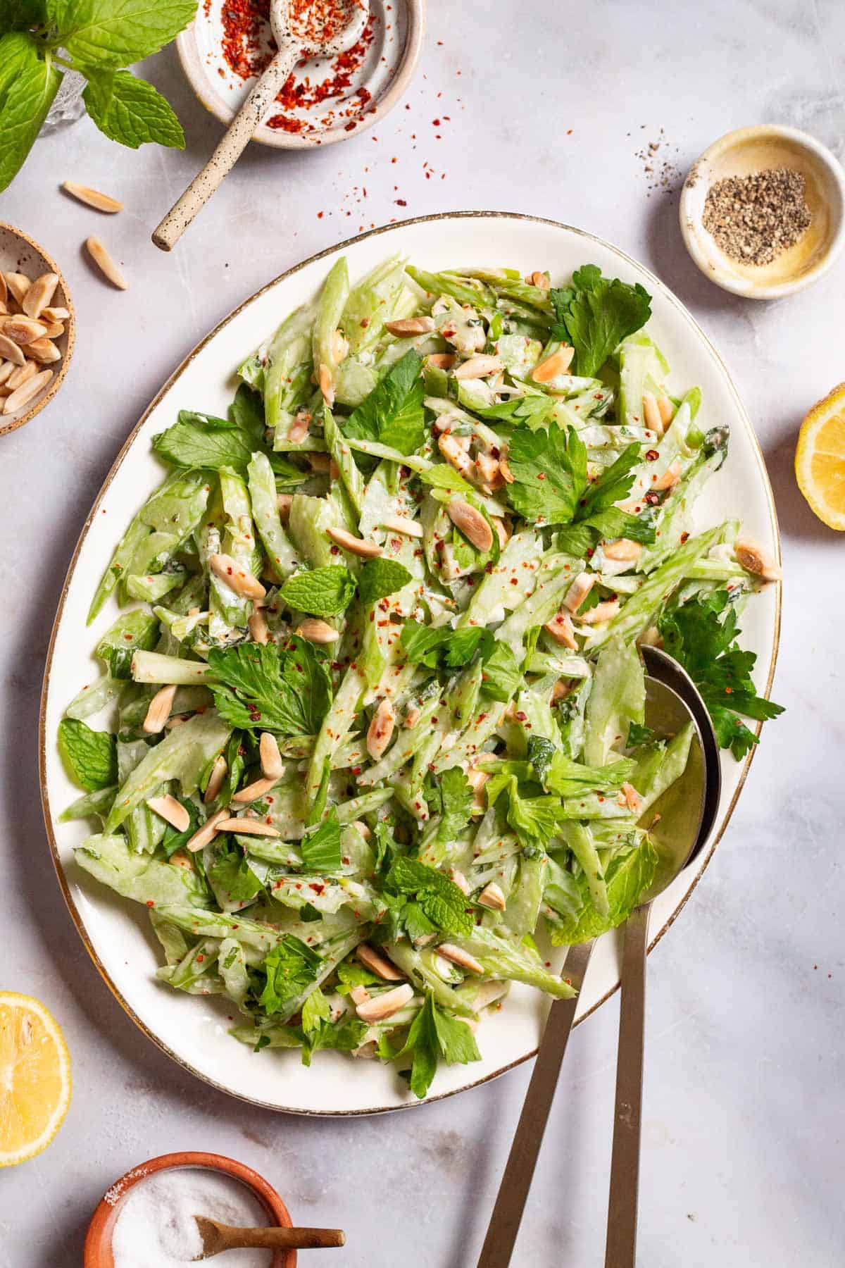 An overhead photo of celery salad on a serving platter with serving utensils. Next to this are bowls of slivered almonds, aleppo pepper, black pepper, salt lemon halves, and mint.