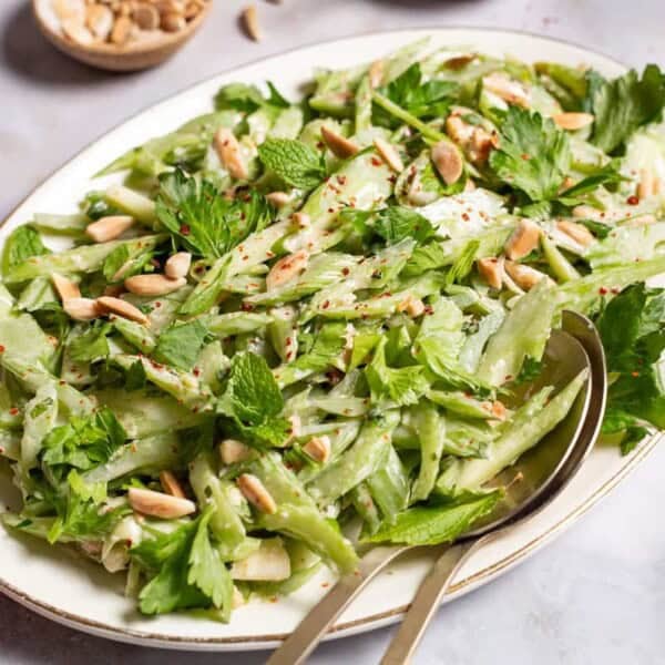 Celery salad on a serving platter with serving utensils. Next to this are bowls of slivered almonds, black pepper, salt and a lemon half.