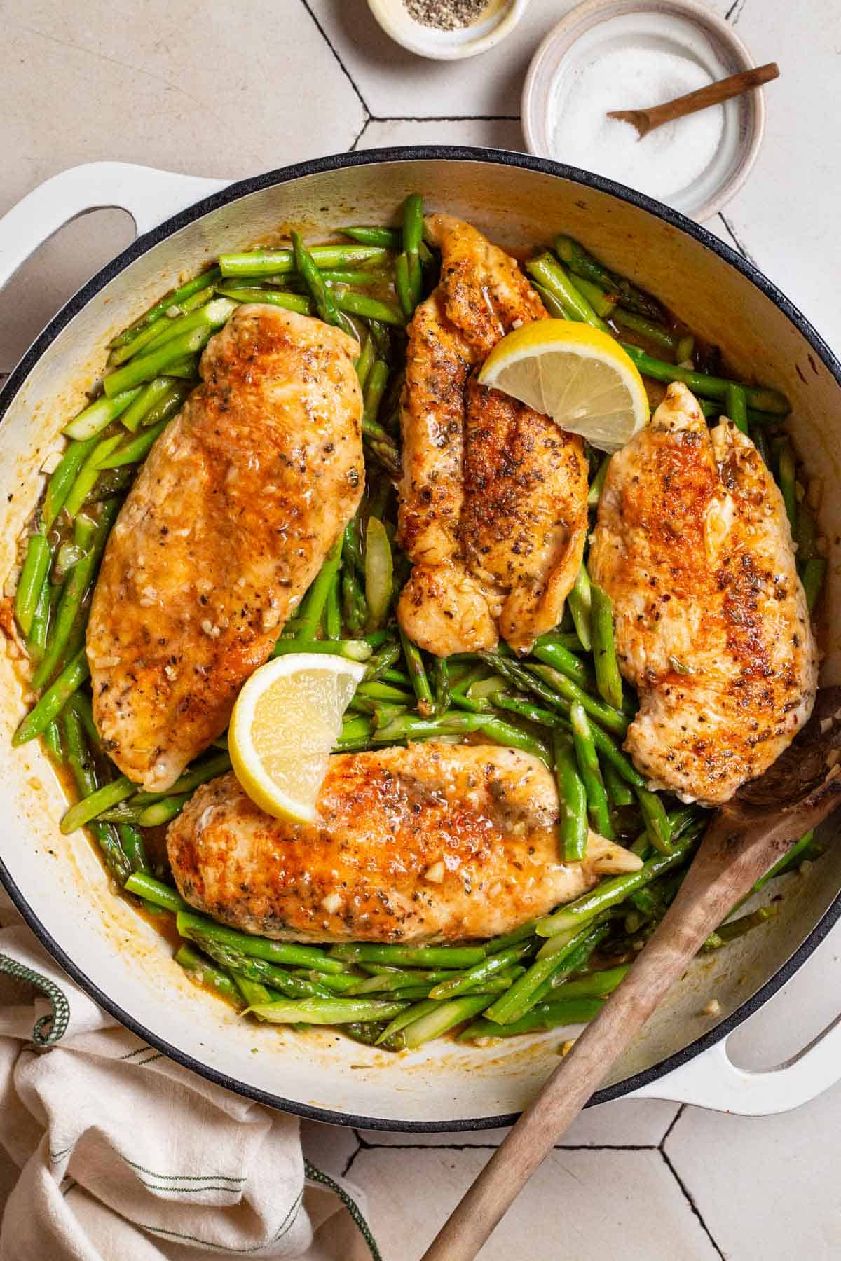 An overhead photo of chicken and asparagus in a skillet with lemon wedges and a wooden spoon. Next to this are small bowls of salt and pepper, and a kitchen towel.