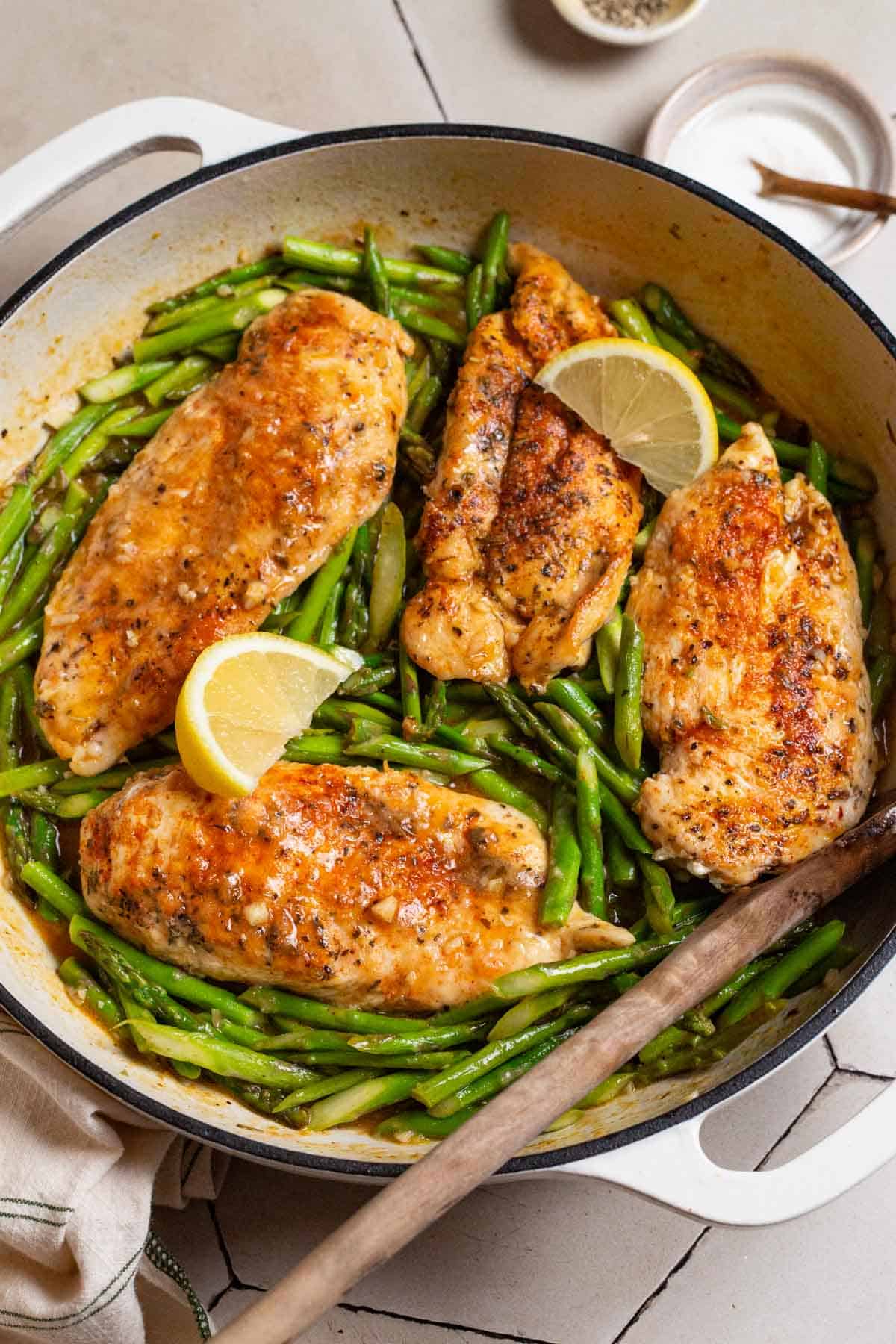 Chicken and asparagus in a skillet with lemon wedges and a wooden spoon. Next to this are two small bowls with salt and pepper.