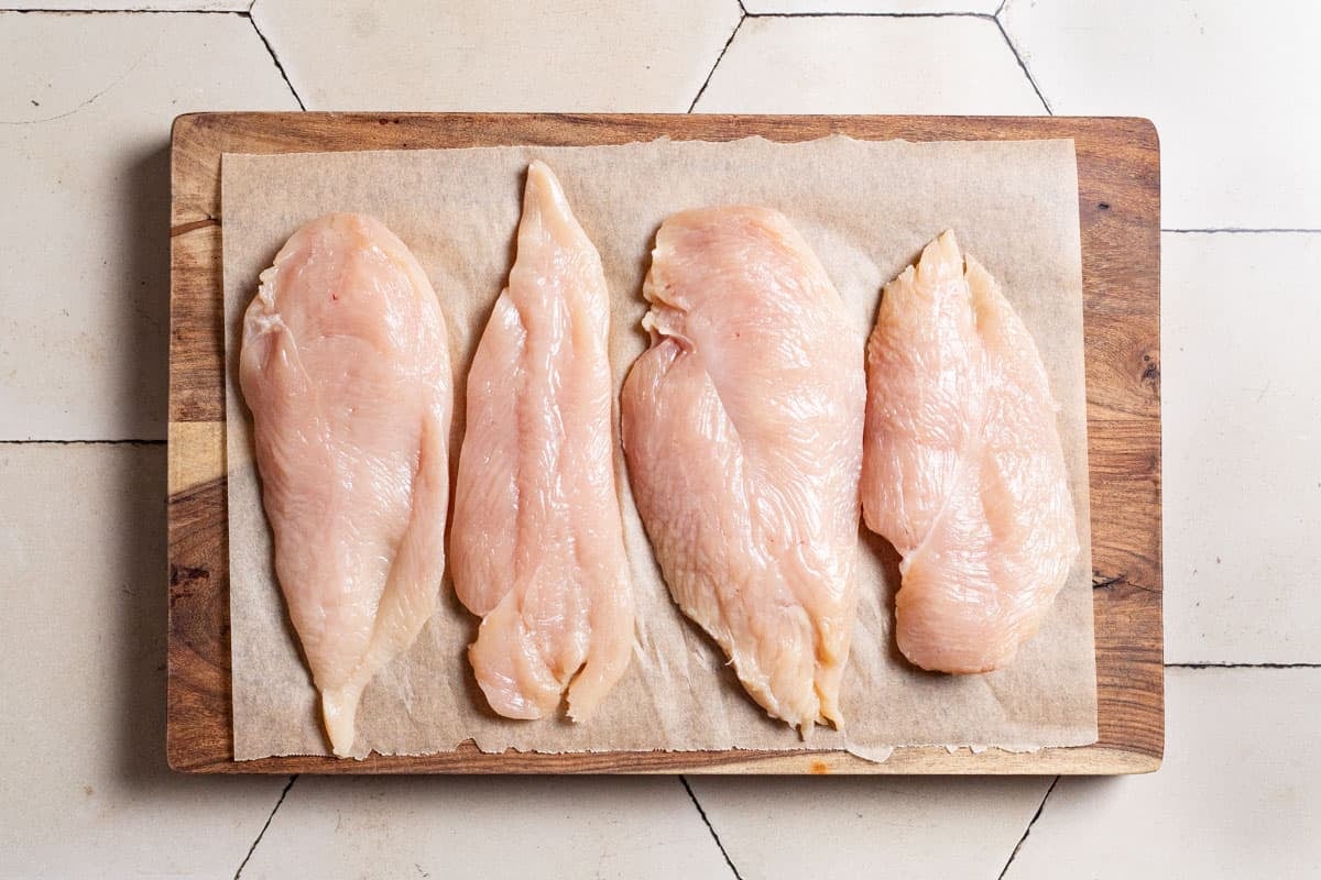 4 uncooked chicken breasts on a parchment lined cutting board.
