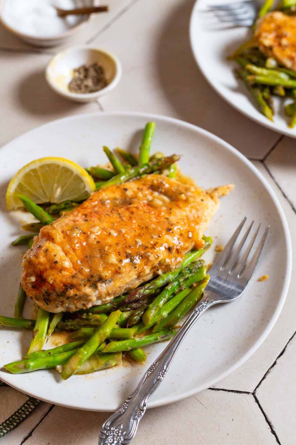 A serving of chicken and asparagus on a plate with a lemon wedge and a fork. In the background, there is another plate of the chicken and asparagus and bowls of salt and pepper.