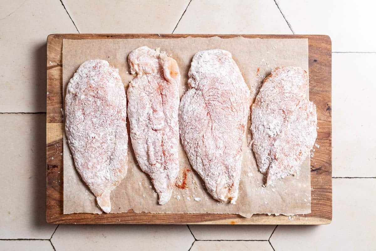 4 seasoned, uncooked chicken breasts coated in flour on a parchment lined cutting board..