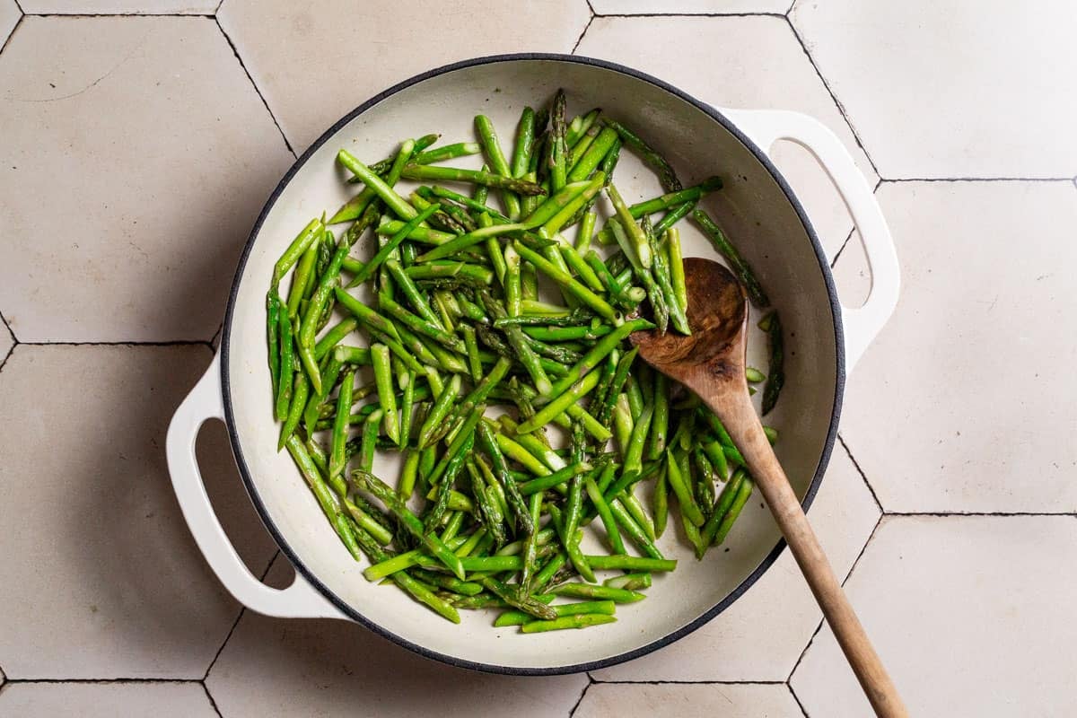 Asparagus pieces being cooked in a skillet with a wooden spoon.