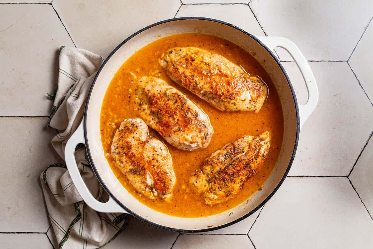 4 Chicken breasts simmering in sauce in a skillet next to a kitchen towel.