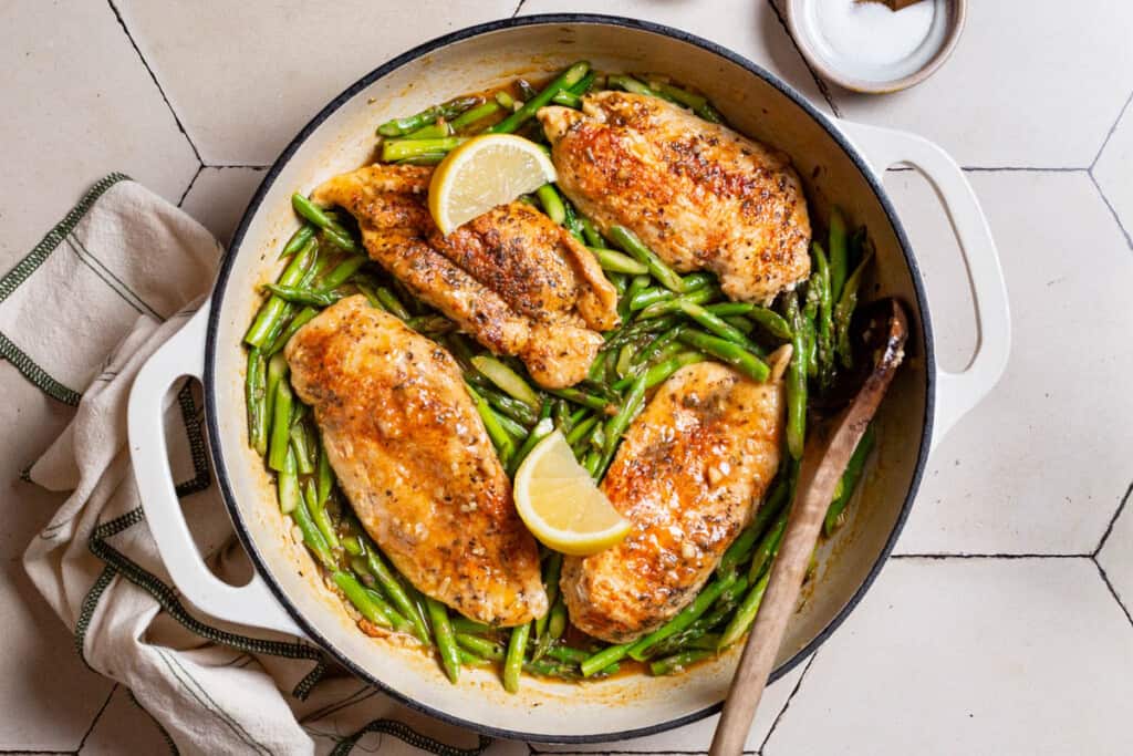 An overhead photo of chicken and asparagus in a skillet with lemon wedges and a wooden spoon. Next to this is a kitchen towel and a small bowl of salt.