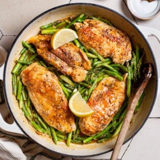 An overhead photo of chicken and asparagus in a skillet with lemon wedges and a wooden spoon. Next to this is a kitchen towel and a small bowl of salt.
